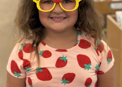 Child wearing yellow LEGO glasses and a dress with strawberry patterns, smiling in the library.
