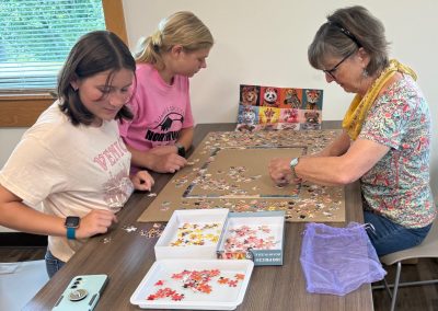 Three people work on a jigsaw puzzle at a table with trays of pieces and a reference image of colorful animals.