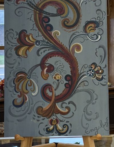The image shows a rectangular wooden panel with an intricate decorative painting displayed on an easel. The panel is predominantly gray with swirling, curved designs in earthy tones of red, gold, brown, and blue. The pattern features ornate, abstract shapes resembling stylized flowers and leaves. These shapes are interconnected by graceful swirls and curls. Some details are enhanced with white highlights and small black dots, adding depth and contrast. The overall style is reminiscent of traditional folk art.