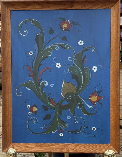 The image depicts a piece of art placed in a wooden frame. The artwork features an intricate floral design on a rich blue background. Prominent swirling green leaves with red accents form the central motif, accompanied by delicate white flowers scattered throughout. The design flows gracefully, with detailed brushstrokes creating a sense of movement and depth.