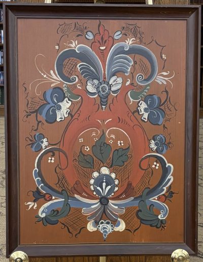 The image is a framed painting featuring symmetrical floral and abstract patterns against a brownish-red background. The composition includes elaborate swirls and curls in shades of blue, white, and gray. The central motif resembles an ornate vase or flower arrangement, with stylized leaves, petals, and a series of circular shapes that provide a sense of balance. The painting is bordered by a dark wooden frame, and it stands on a display tripod. The overall style appears decorative, with intricate detailing and vibrant color contrasts.