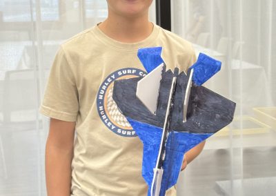 Young person holding a blue and black craft model airplane, wearing a beige T-shirt and a Bass Pro Shops cap.