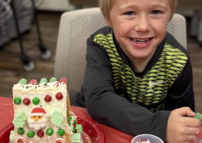 Child smiling next to a decorated gingerbread house on a red plate with colorful candies and sprinkles nearby.