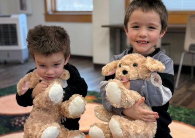 Two children holding stuffed dog toys in a room with wooden floors and large windows.
